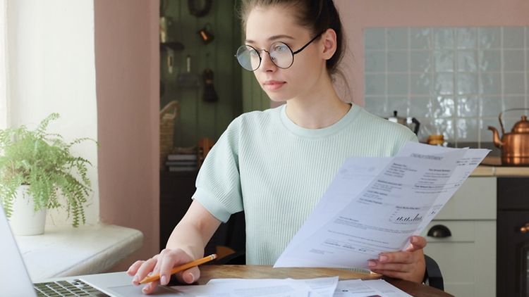  Symbolic image: Young woman managing domestic budget, sitting at kitchen table with open laptop, documents and calculator, using touchpad, making notes with pencil 