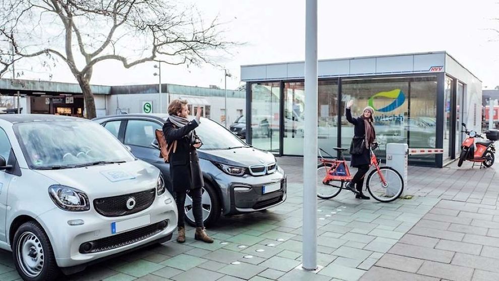 Ein Mann und eine Frau winken sich zu. Die Frau hält ein rotes Hamburger Stadtrad. Der Mann steht zwischen zwei kleinen Autos.