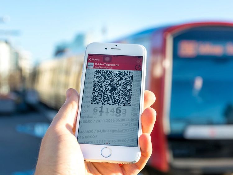  Eine Hand hält ein Smartphone, auf dem ein Ticket mithilfe eines QR-Codes abgebildet ist. Im Hintergrund ist eine S-Bahn verschwommen zu sehen.