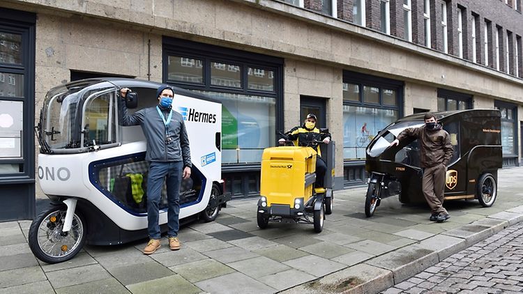 Three persons standing in front of three different types of cargo bikes by Hermes, Deutsche Post and UPS.