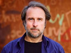  Portrait of actor Bjarne Mädel smirking for the camera. He has dark blonde hair, blue eyes and a shaggy beard, and he is wearing a dark blue shirt over a black t-shirt.