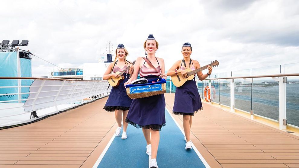 Three women with blue skirts and red and white striped tops hold instruments in their hands and walk along a ship. 