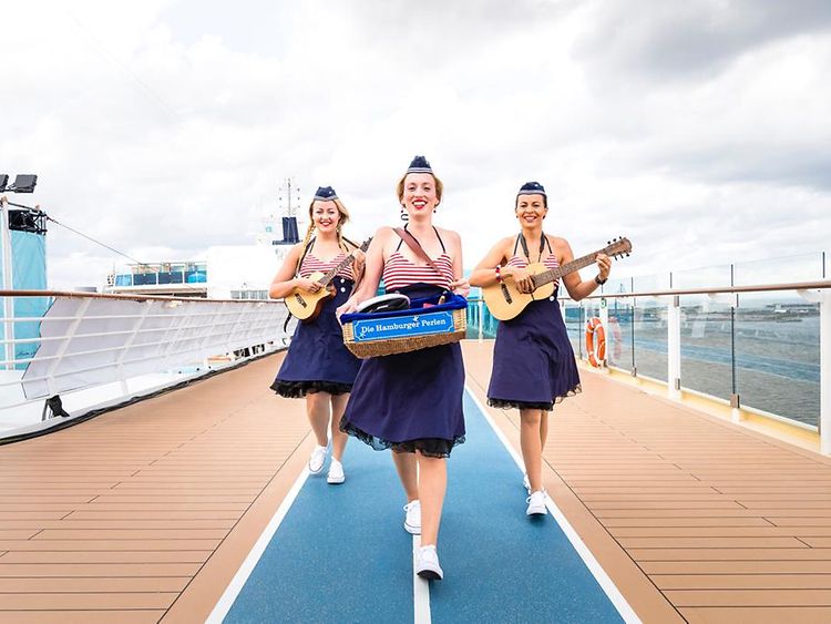  Three women with blue skirts and red and white striped tops hold instruments in their hands and walk along a ship. 