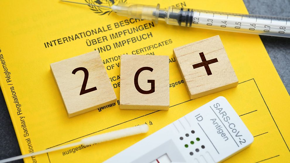 A yellow German book of vaccinations with syringe, testing equipment and the letters 2, G and a plus sign on top of it.