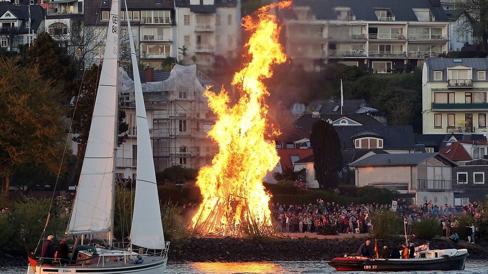 An Easter bonfire close to the Elbe river in Hamburg-Blankenese, with crowd gathering and sail boat and bark in front