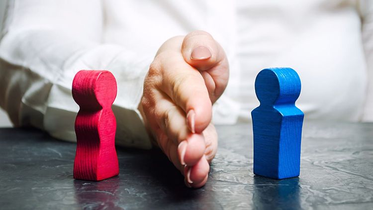  Eine Hand trennt zwei auf einem Tisch stehende Holzfiguren. Die eine Figur ist rot, die andere blau.