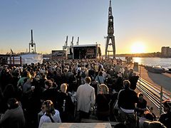 A crowd of people in front of a live-music stage with sunset against Hamburg's harbour front