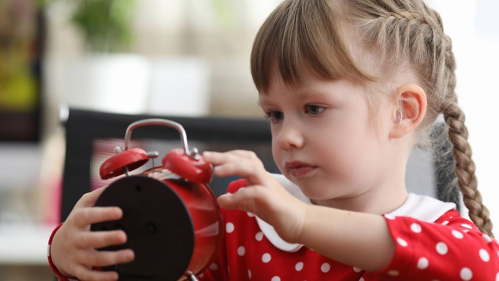  Little girl with pigtails looking at red alarm clock at table