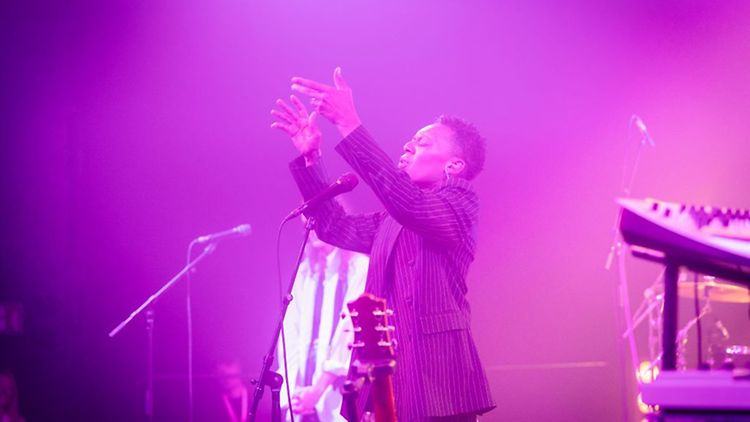  Eine Person steht auf einer Bühne, hebt die Arme und singt in ein Mikrofon. Die Bühne ist in lila Licht getaucht.