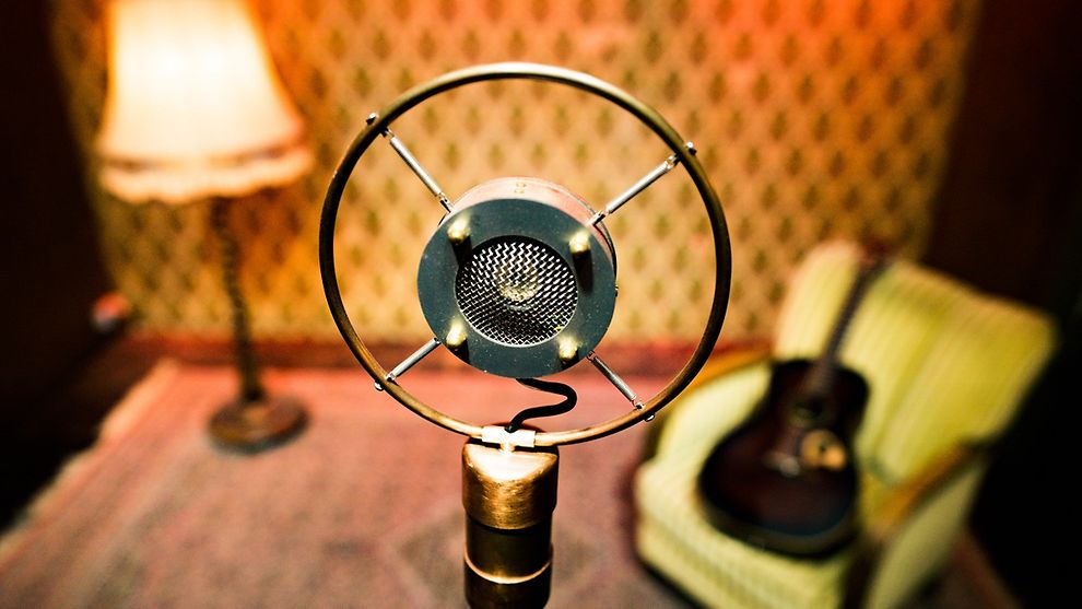 Vintage microphone on a stand on a stage with vintage lamp and acoustic guitar on vintage armchair in background