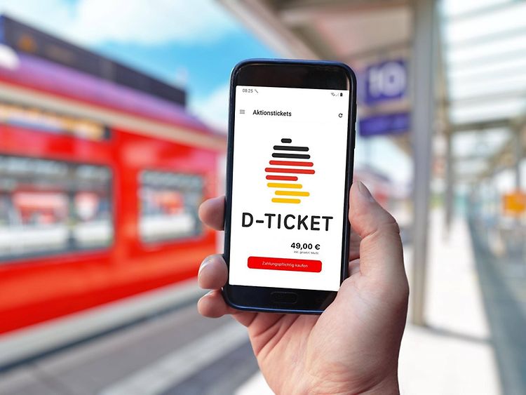  A hand holding a smartphone with the Deutschlandticket on display with train station in background