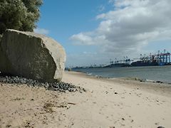  A giant boulder sitting on a sand beach by the river; people walking by; port cranes in background