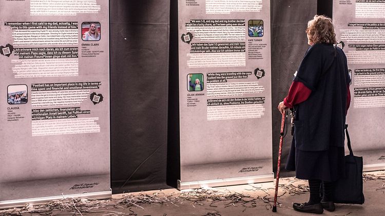 Eine ältere Frau steht vor Ausstellungsplakaten des Vereins Football Supporters Europe e.V..
